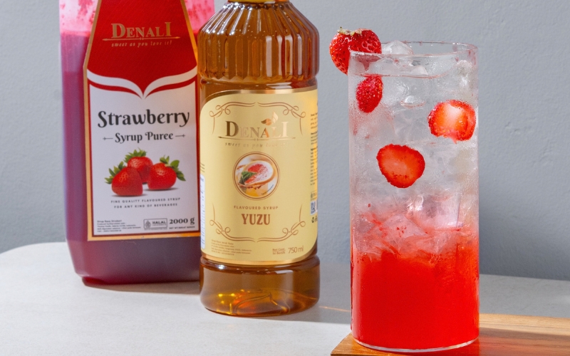 Mocktail Berry Blossom bergradasi merah marun ke keemasan dalam gelas highball bergelembung soda, dihiasi buah stroberi segar dan olesan selai strawberry.
